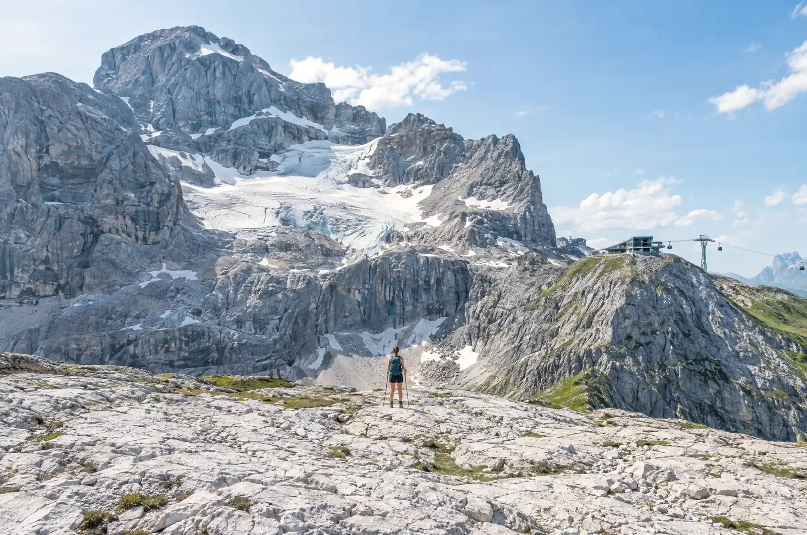 Hiker on a limestone plateau below a rock face with a glacier and a cable car top station
